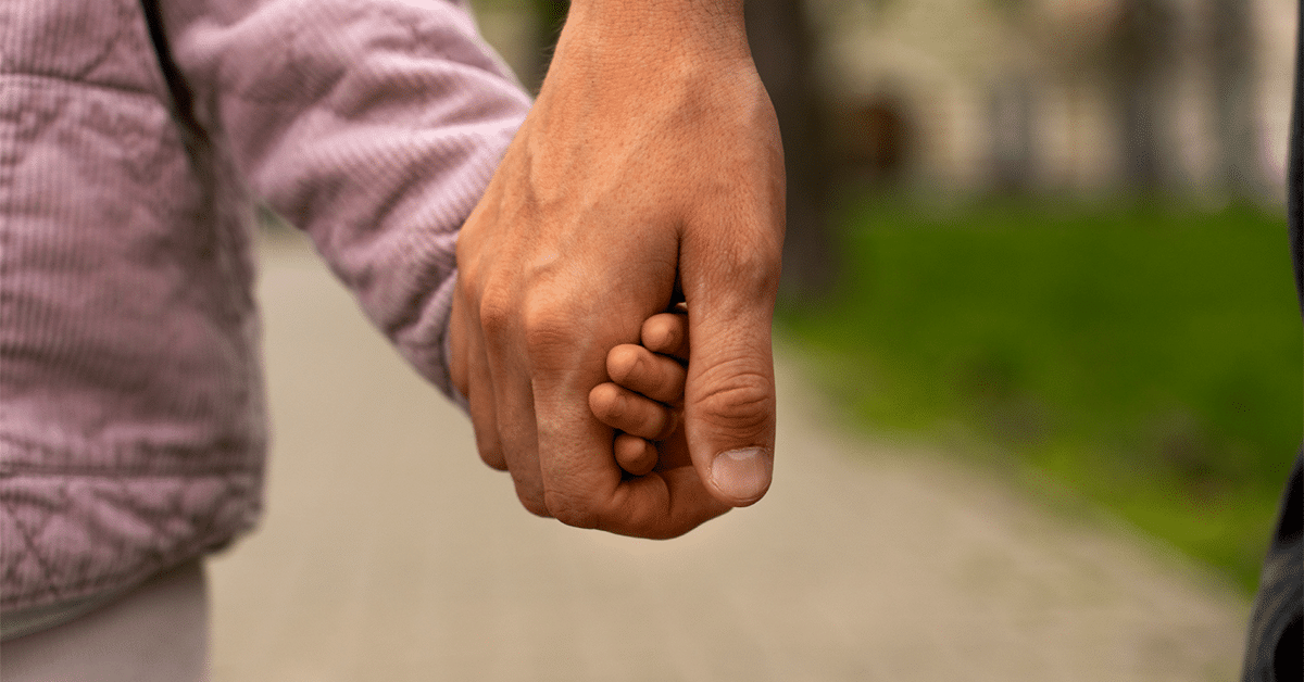 Child Support–A close-up of a father holding his daughter's hand as they walk together outside.