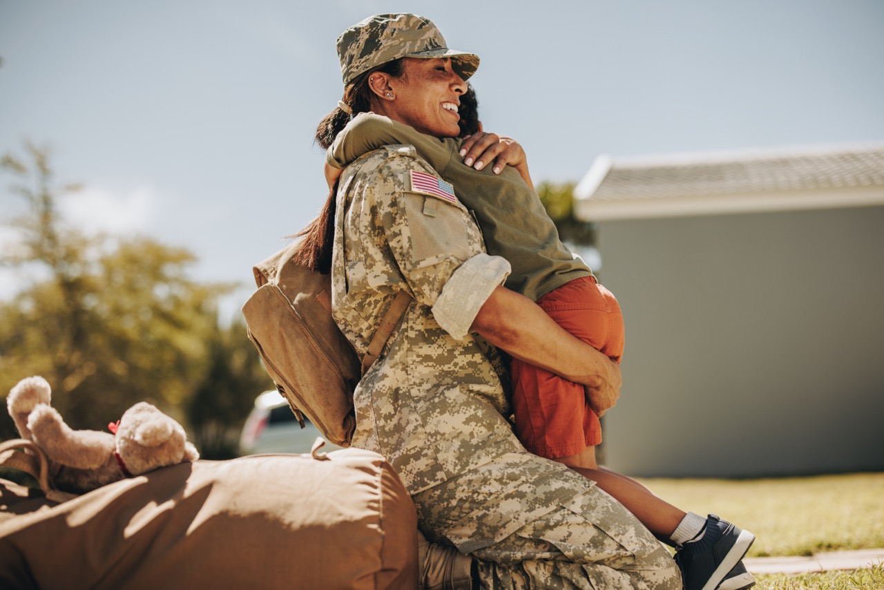 A female soldier in camouflage uniform, carrying a backpack, joyfully hugging her child outdoors. The child, dressed in a green shirt and orange shorts, is lifted into the air as they embrace. A stuffed animal is visible on the ground near a duffel bag in the foreground, with a home and trees blurred in the background.