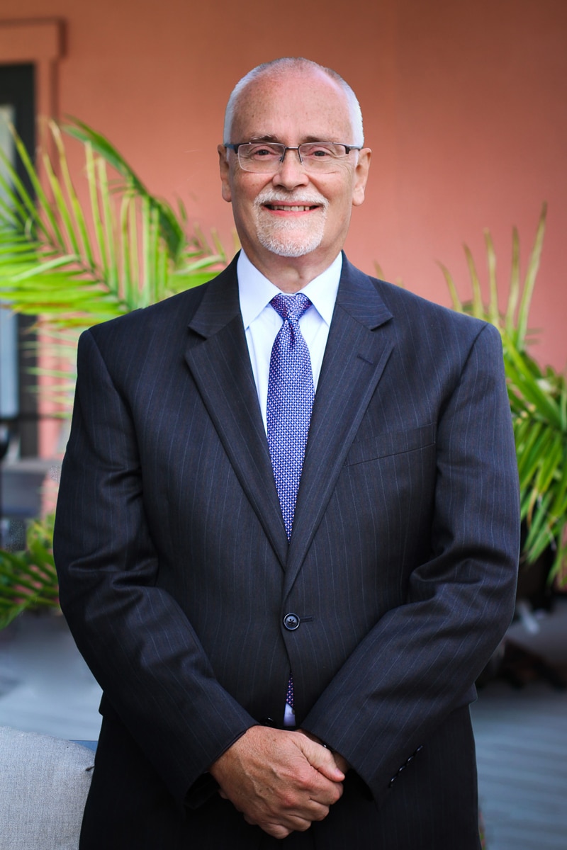 Man standing with hands crossed in the front, waring a dark blue suit and glasses, smiling at the camera, lawyer