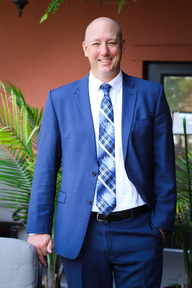 Tall man wearing blue suit and blue plaid tie, standing outside and smiling at the camera, lawyer headshot